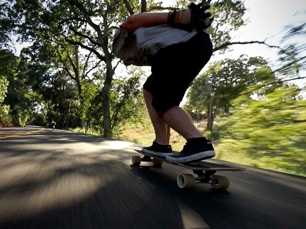 Longboarder in a tuck bombing down a tree-lined road on Avenue suspension trucks
